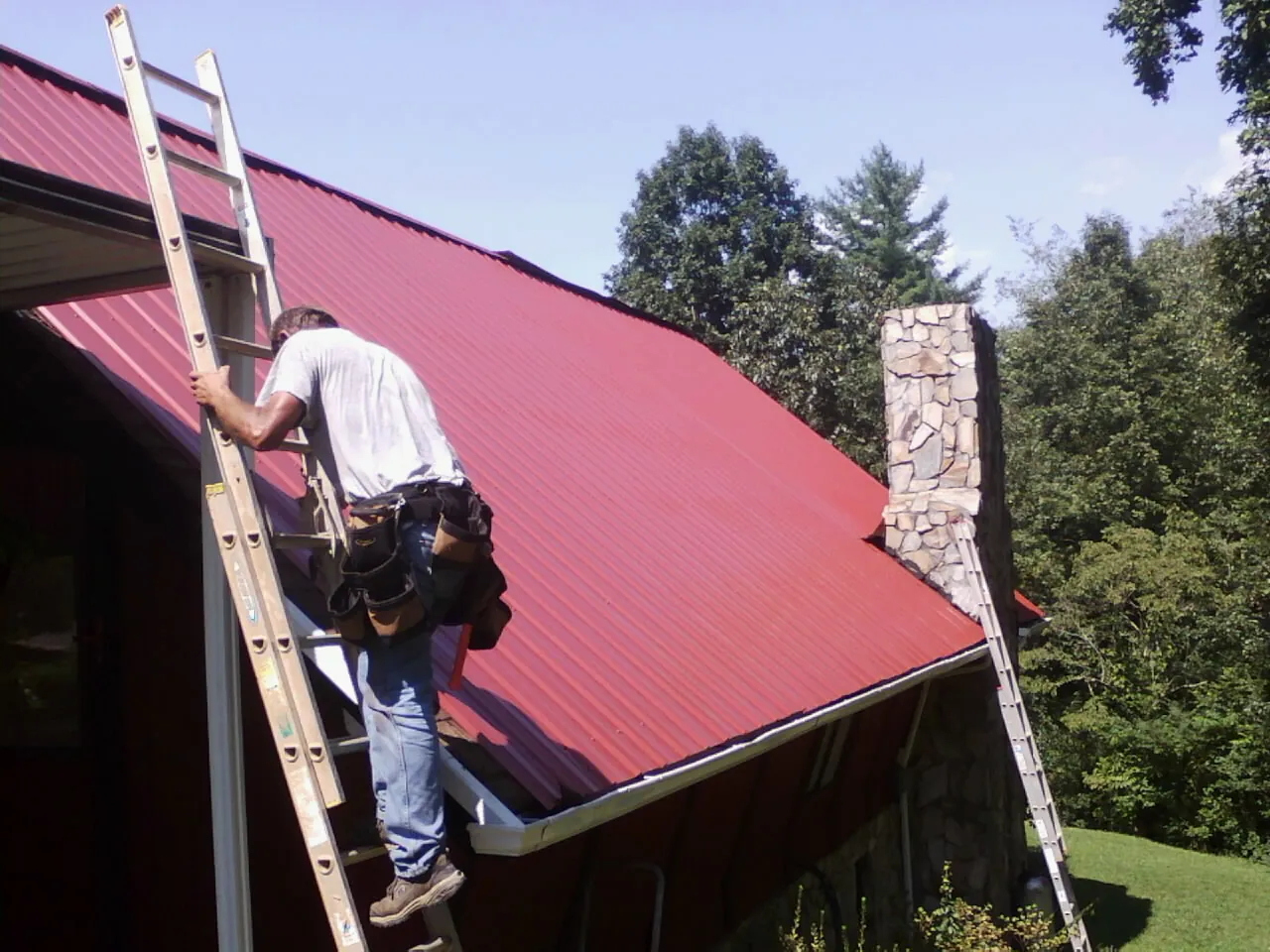 Roofer on a ladder beside a red metal roof and stone chimney — Soffit & Fascia Repair by Roofing Brothers in Punxsutawney